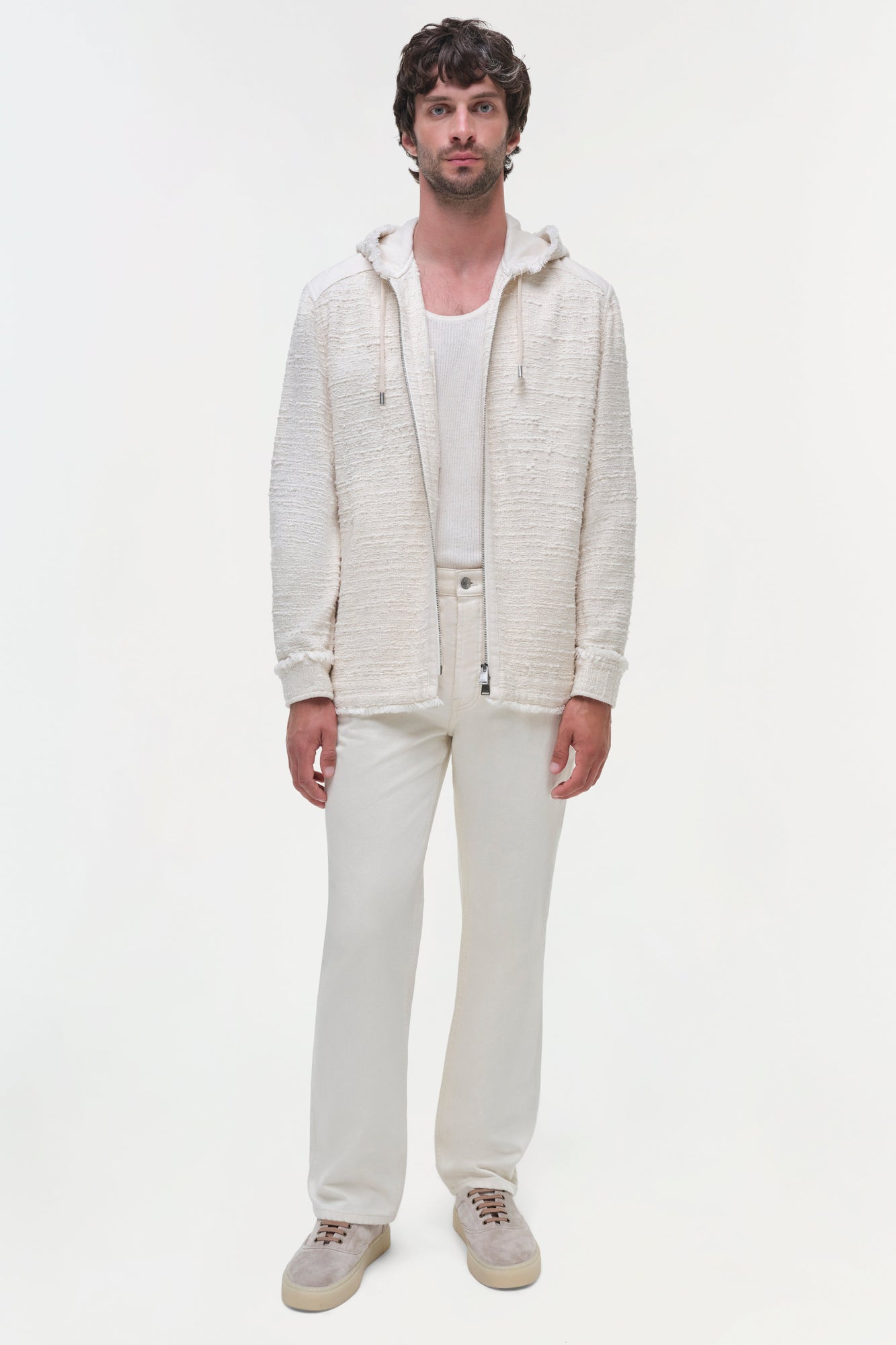 Man wearing cream zip-up hoodie jacket over white ribbed shirt, paired with cream pants and suede sneakers.