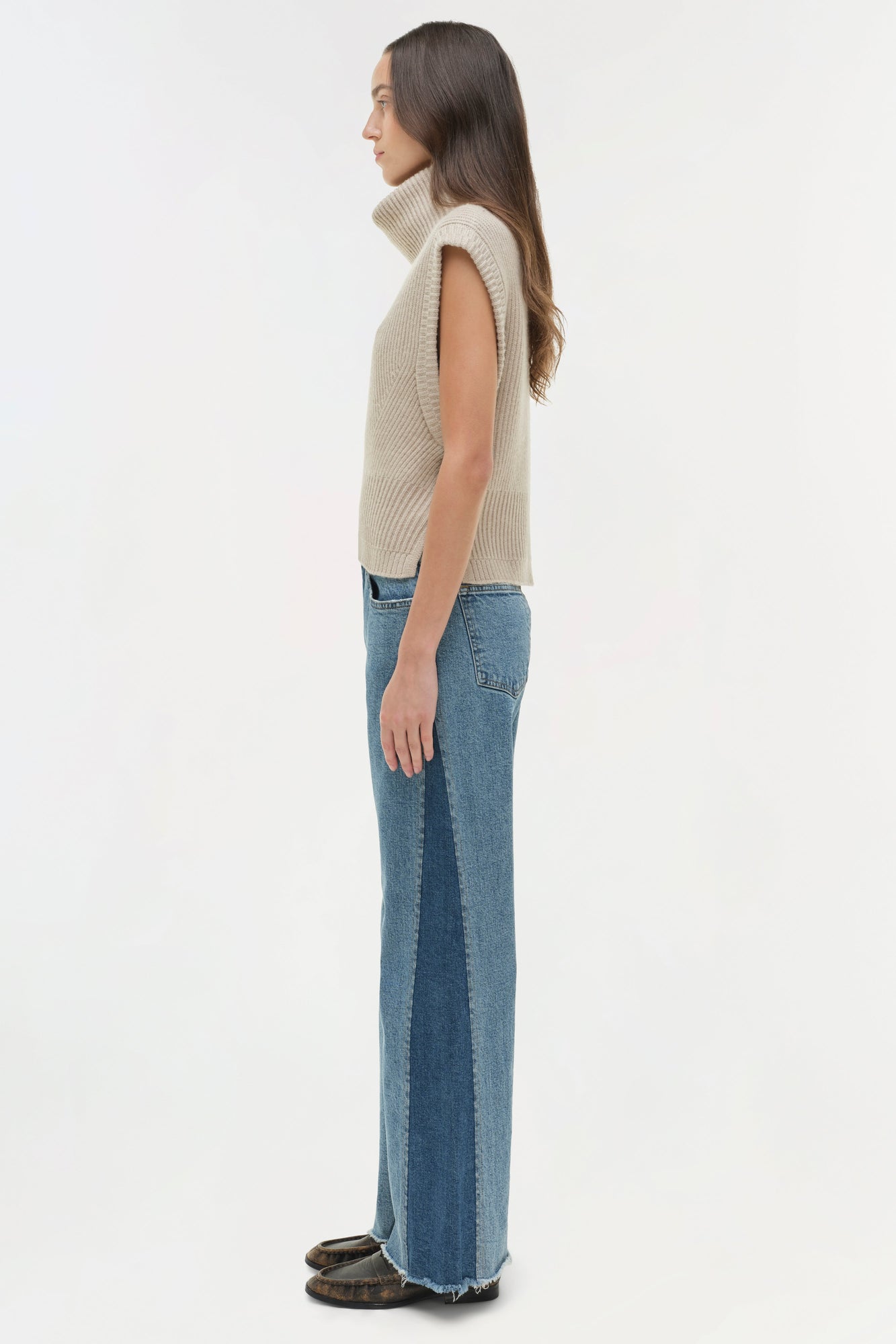 Woman wearing cream sleeveless turtleneck and blue straight leg jeans, side view.