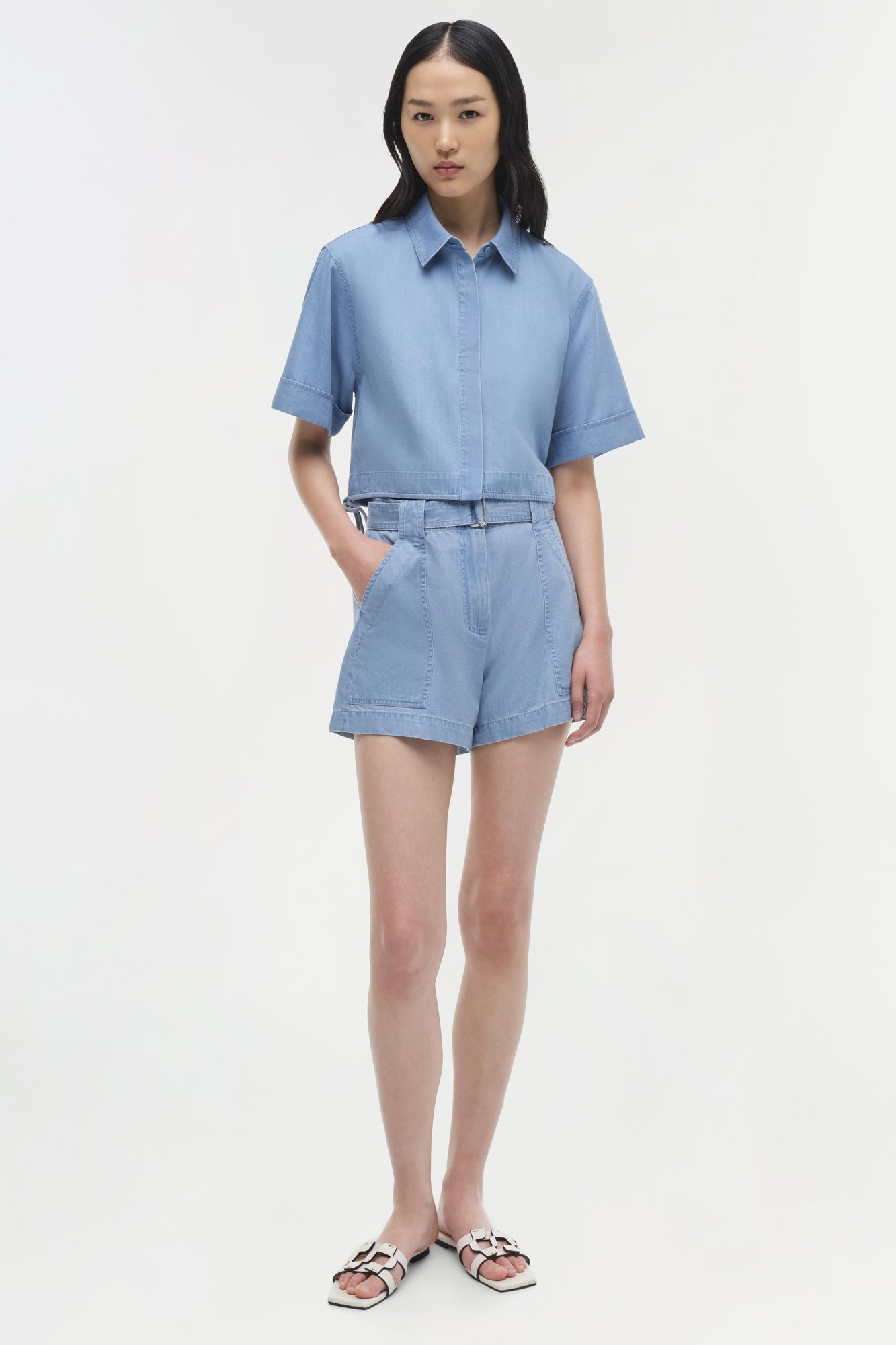 Light blue denim button-up shirt with short sleeves and cropped length, styled with matching denim shorts.