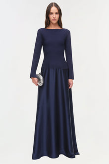 Woman wearing navy satin knit gown with long sleeves, fitted bodice, and flowing skirt to floor, holding metallic clutch.