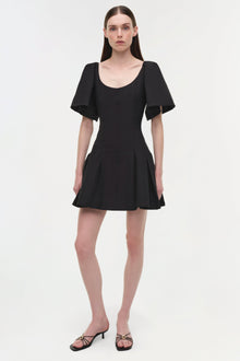 Black mini dress with short puffed sleeves, scoop neckline, and pleated skirt detail worn with black strappy heels.