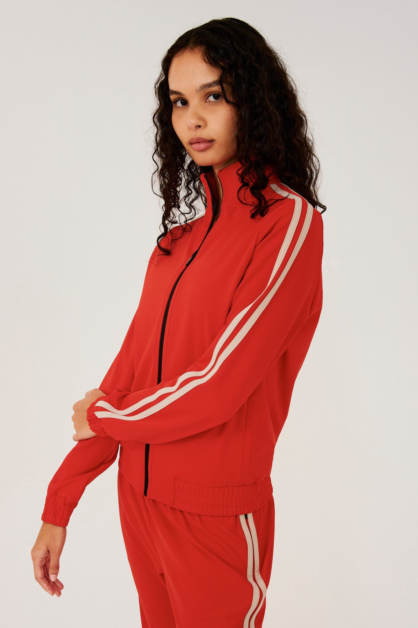 Side view of girl wearing red zip jacket that stops under chin with two white stripes down the side and a black zipper in the front with a red sweatpants