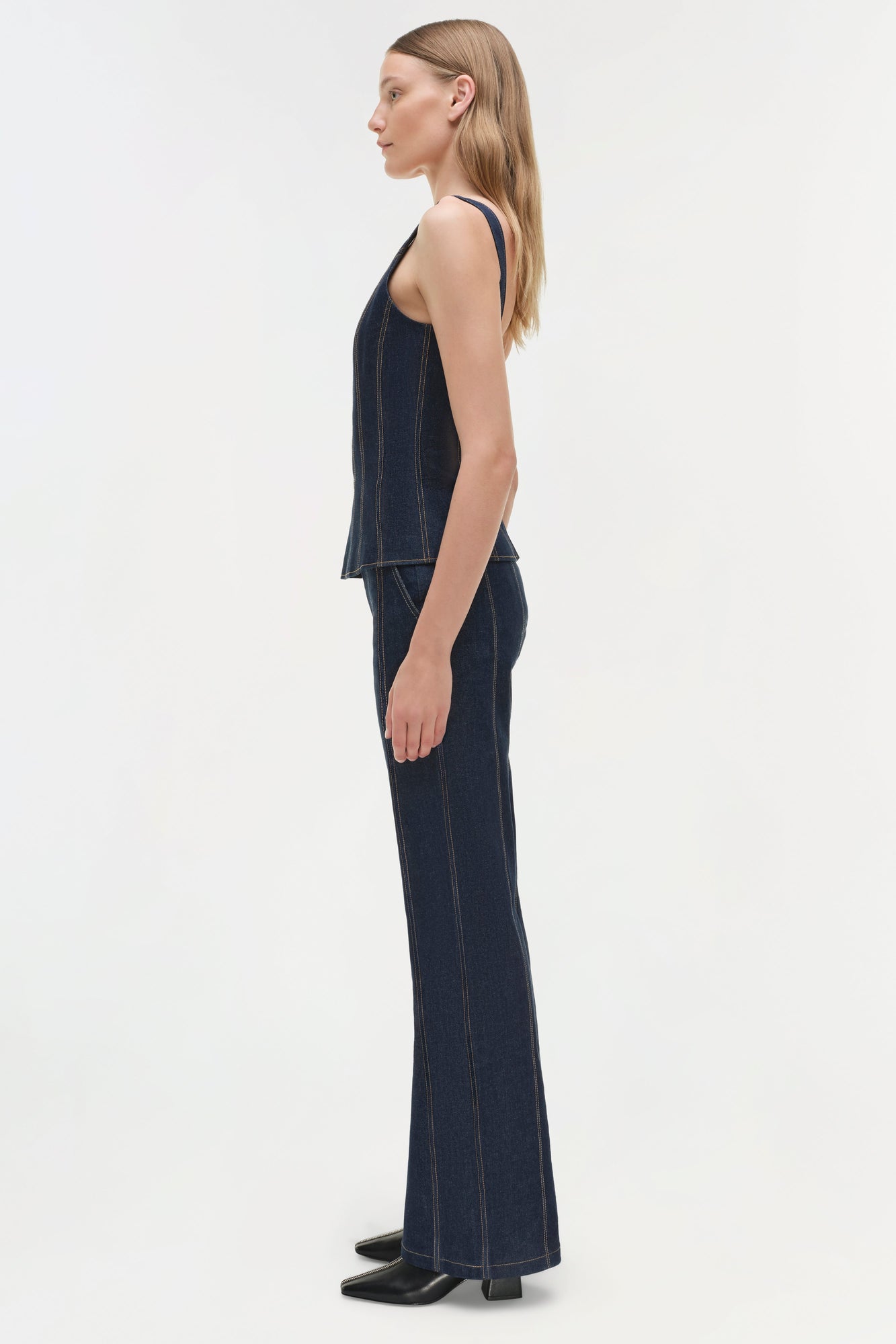 Woman wearing dark indigo denim trousers with visible seaming and a sleeveless denim top, shown from the side.