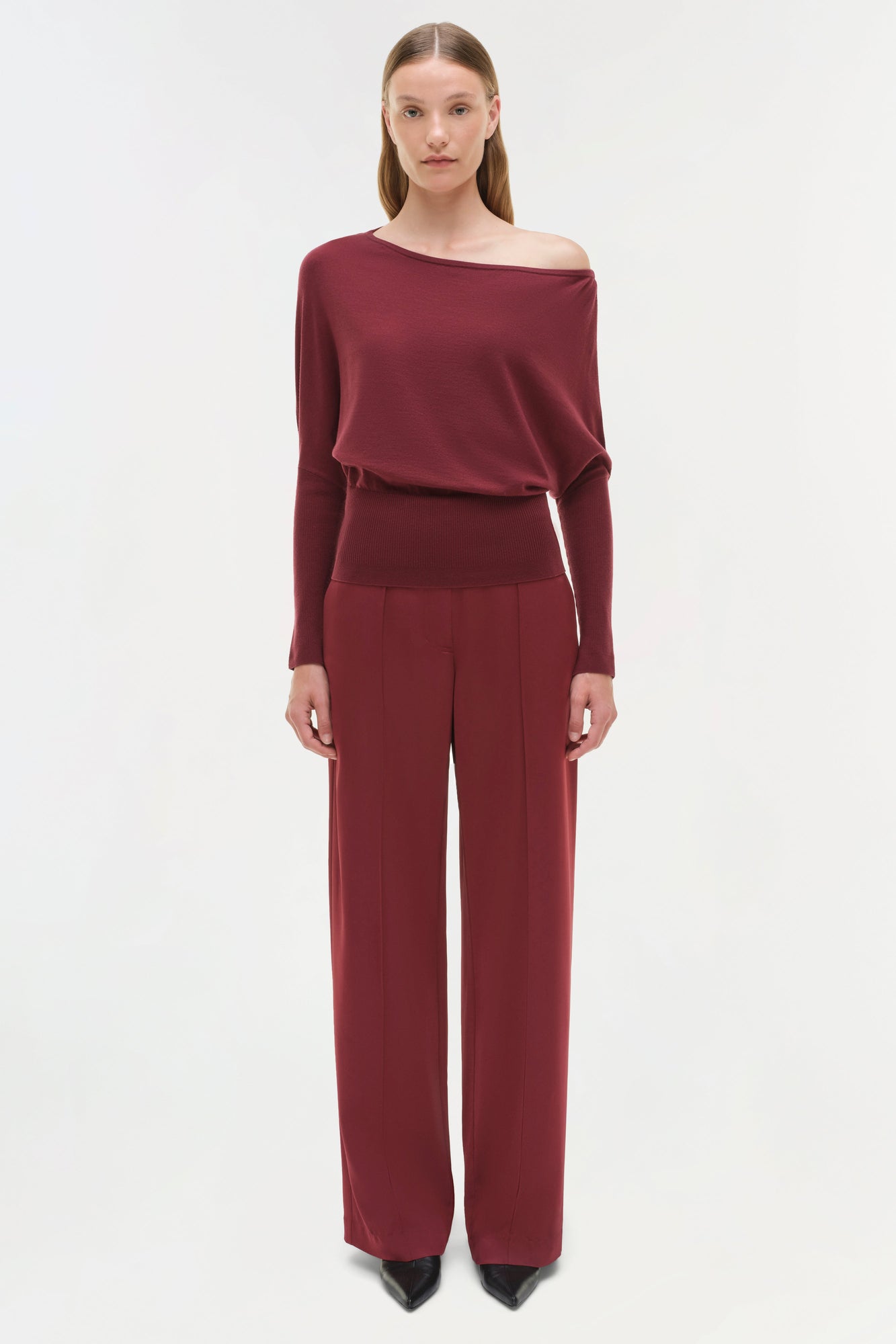 Woman wearing burgundy satin wide-leg pants with a matching off-shoulder long-sleeve top and black pointed-toe boots.