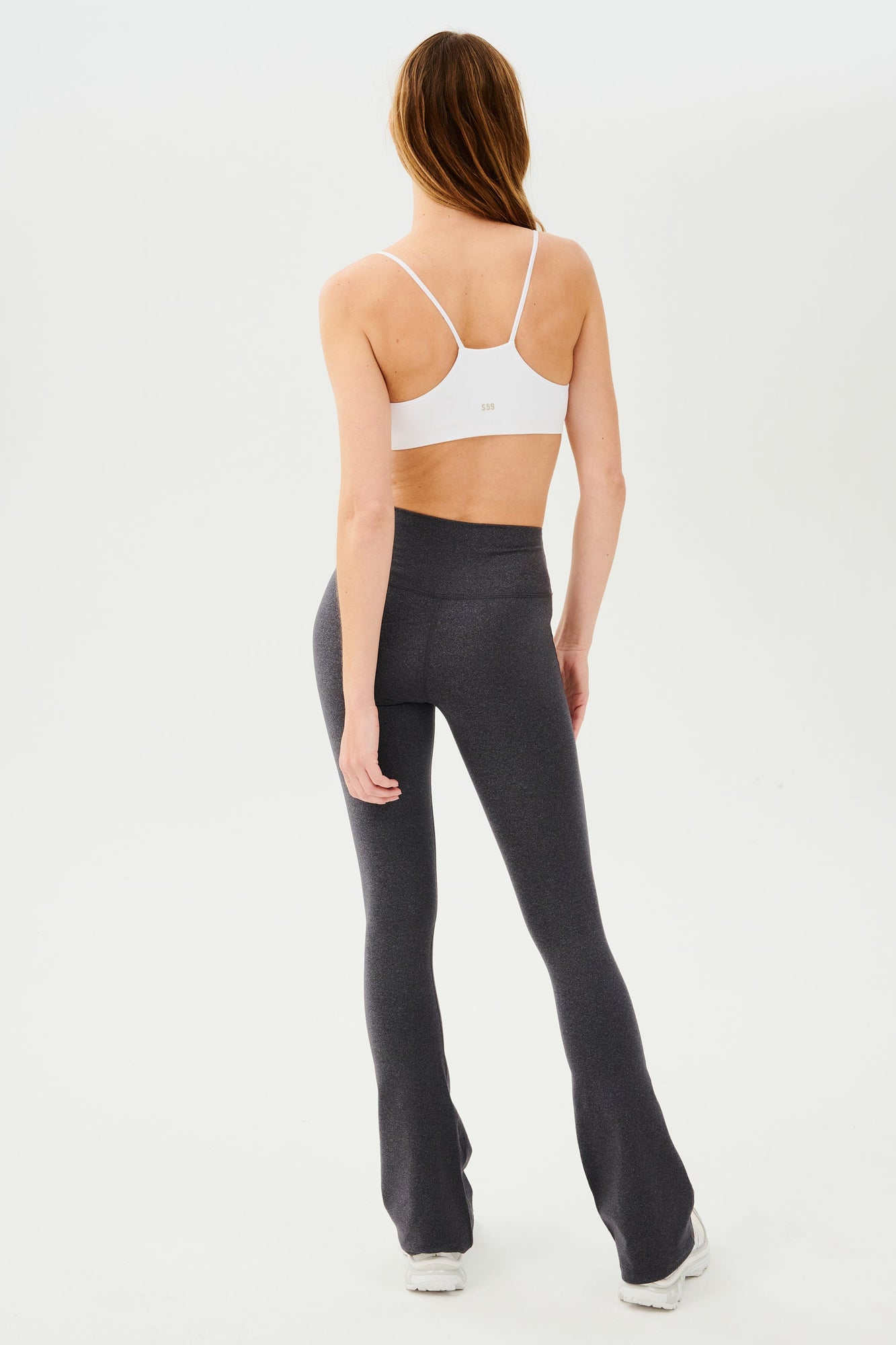 Full back view of woman with straight blonde hair wearing dark gray high waist below ankle length legging with wide flared bottoms and white racerback bra with spaghetti straps. Paired with white shoes.