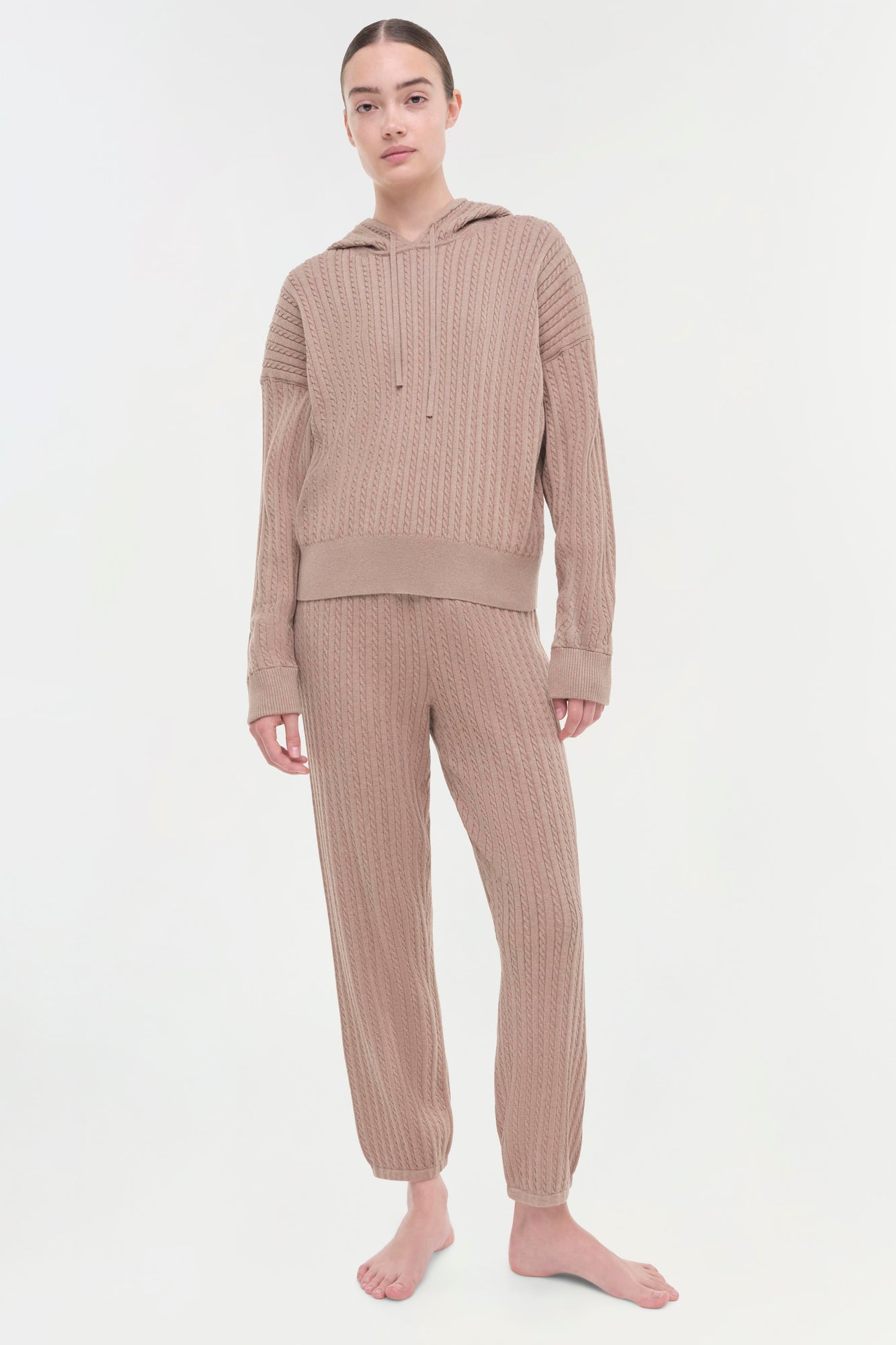 Beige cable knit hoodie and matching pants set on model against white background.