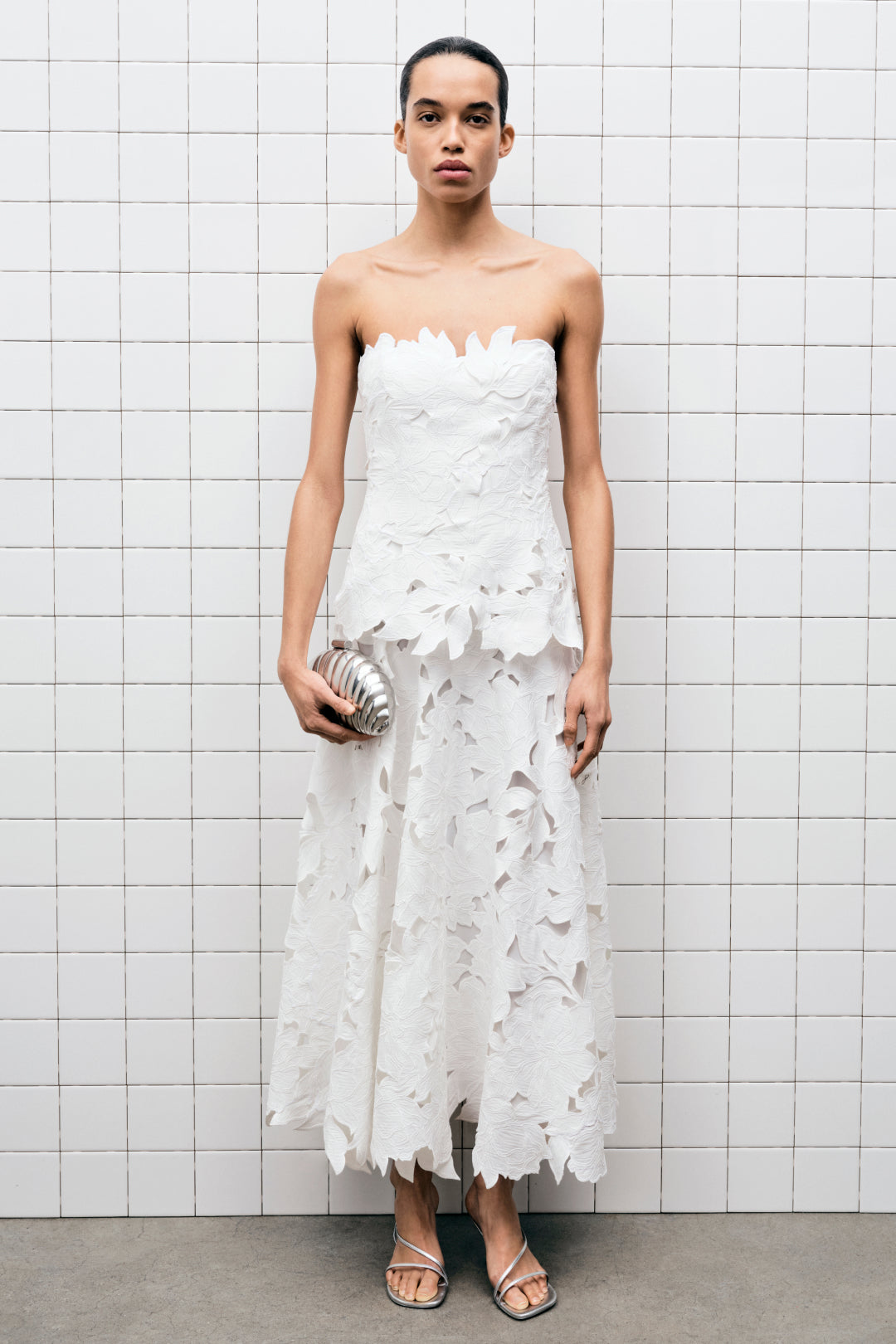 White strapless midi dress with leaf-shaped embroidered cutouts throughout, worn with metallic sandals.