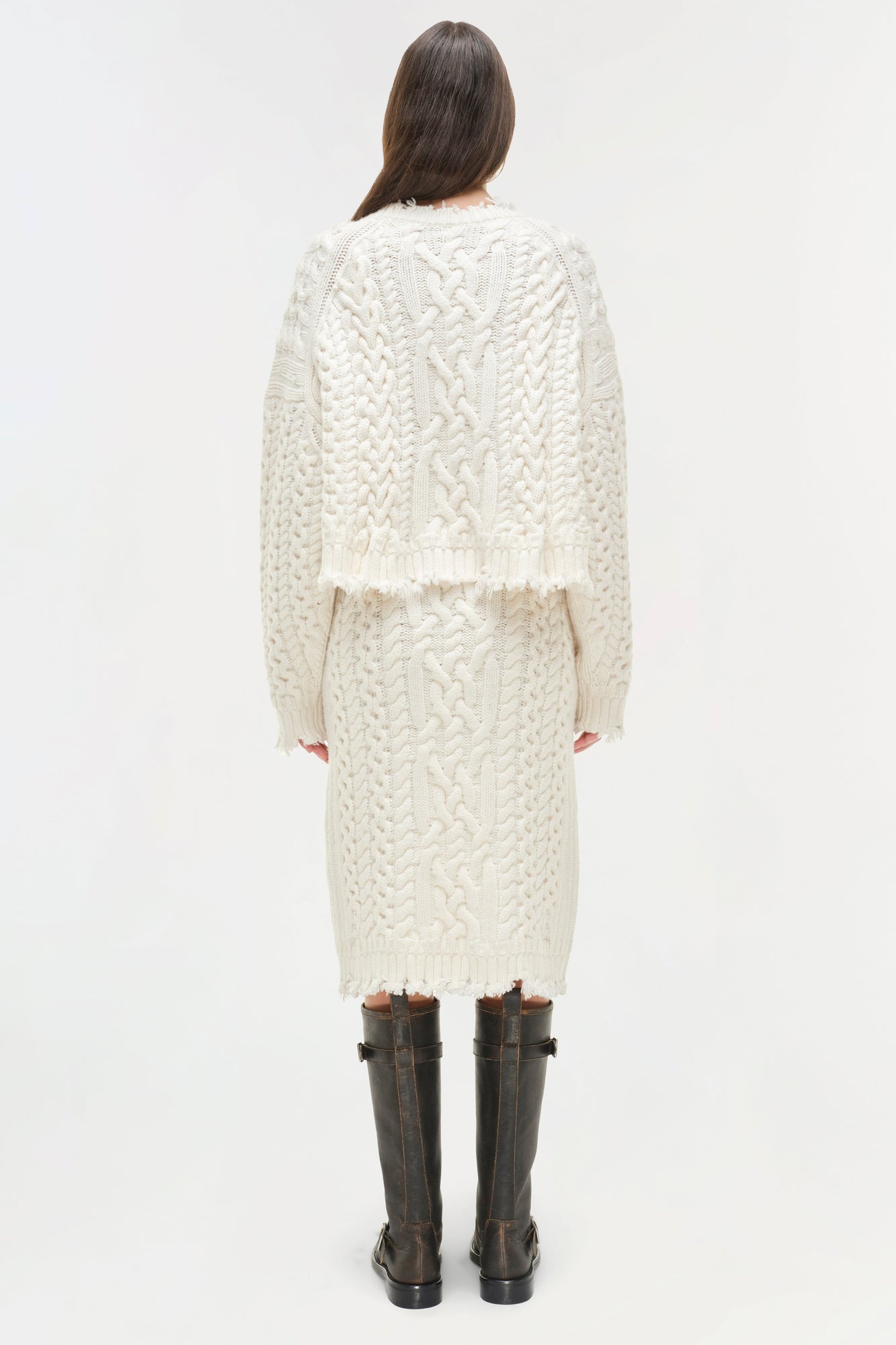 Cream cable-knit midi skirt with distressed hem, worn with matching cropped cardigan and black boots.