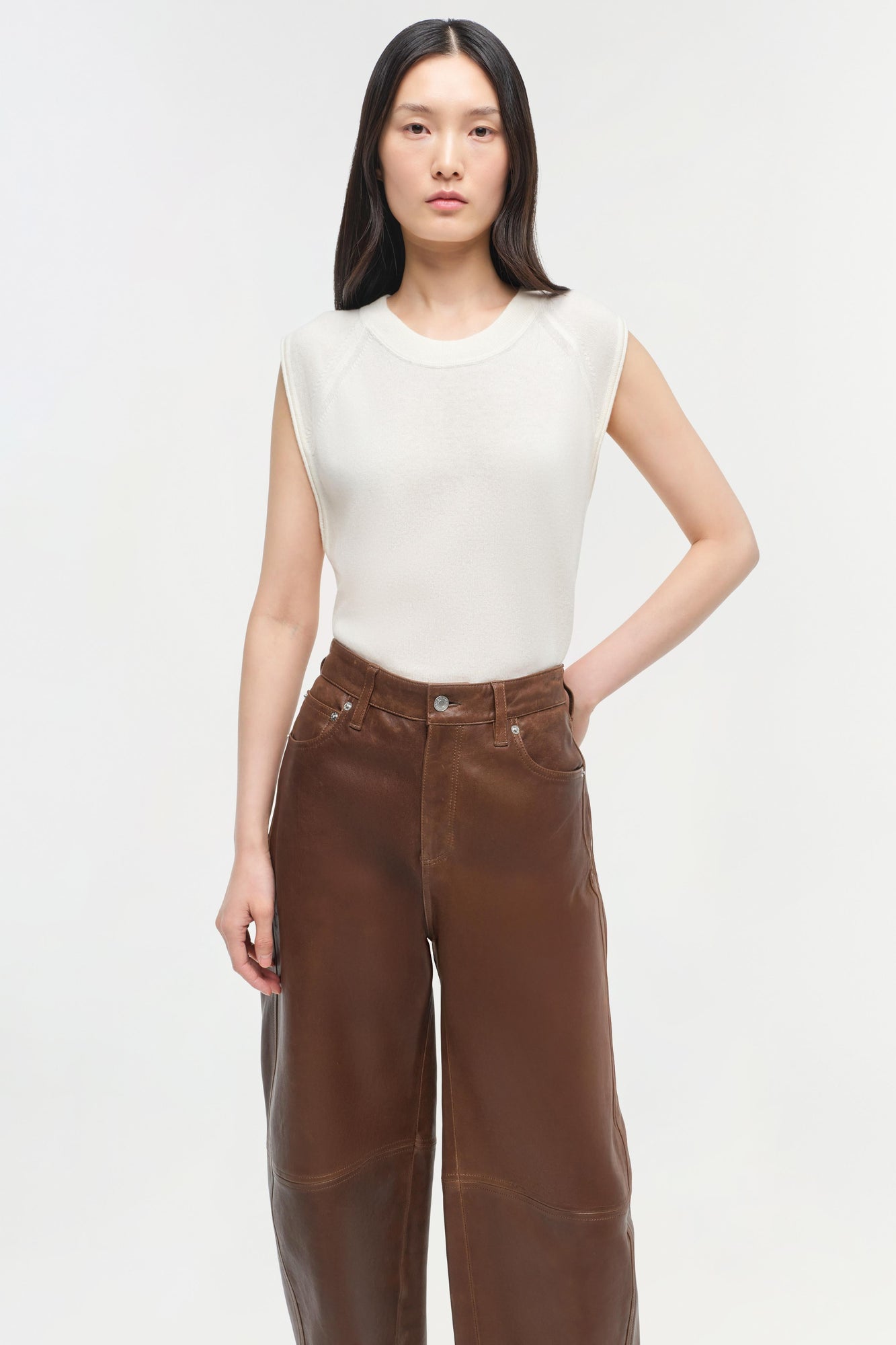 Woman wearing cream sleeveless cashmere vest with brown leather pants against white background.