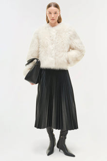 Woman wearing cream-colored shearling jacket with black pleated skirt and black boots, holding a black handbag.
