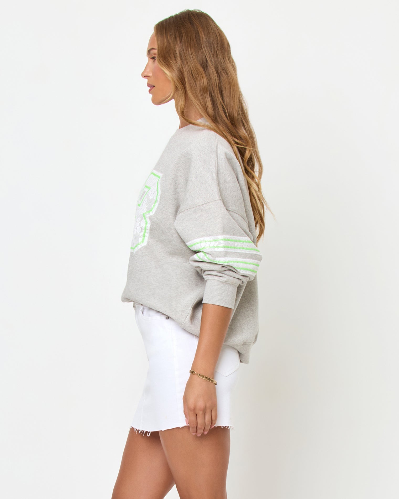 Side three-quarter studio view of the Legend Sweatshirt in Limeade, a heather gray oversized crewneck sweatshirt with a neon green graphic, styled with a white denim skirt.