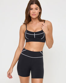 Close-up front view from the knees up of the LSPACE Eclipse Bra in black on a model against a white background, showing the bra's trim detail, paired with matching black-cream active shorts.