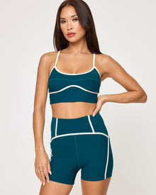 Close-up front view of model wearing the Revel Top in deep teal-cream showing the scoop neckline and cream piping detail, with matching deep teal-cream Revel Short, hand on hip.