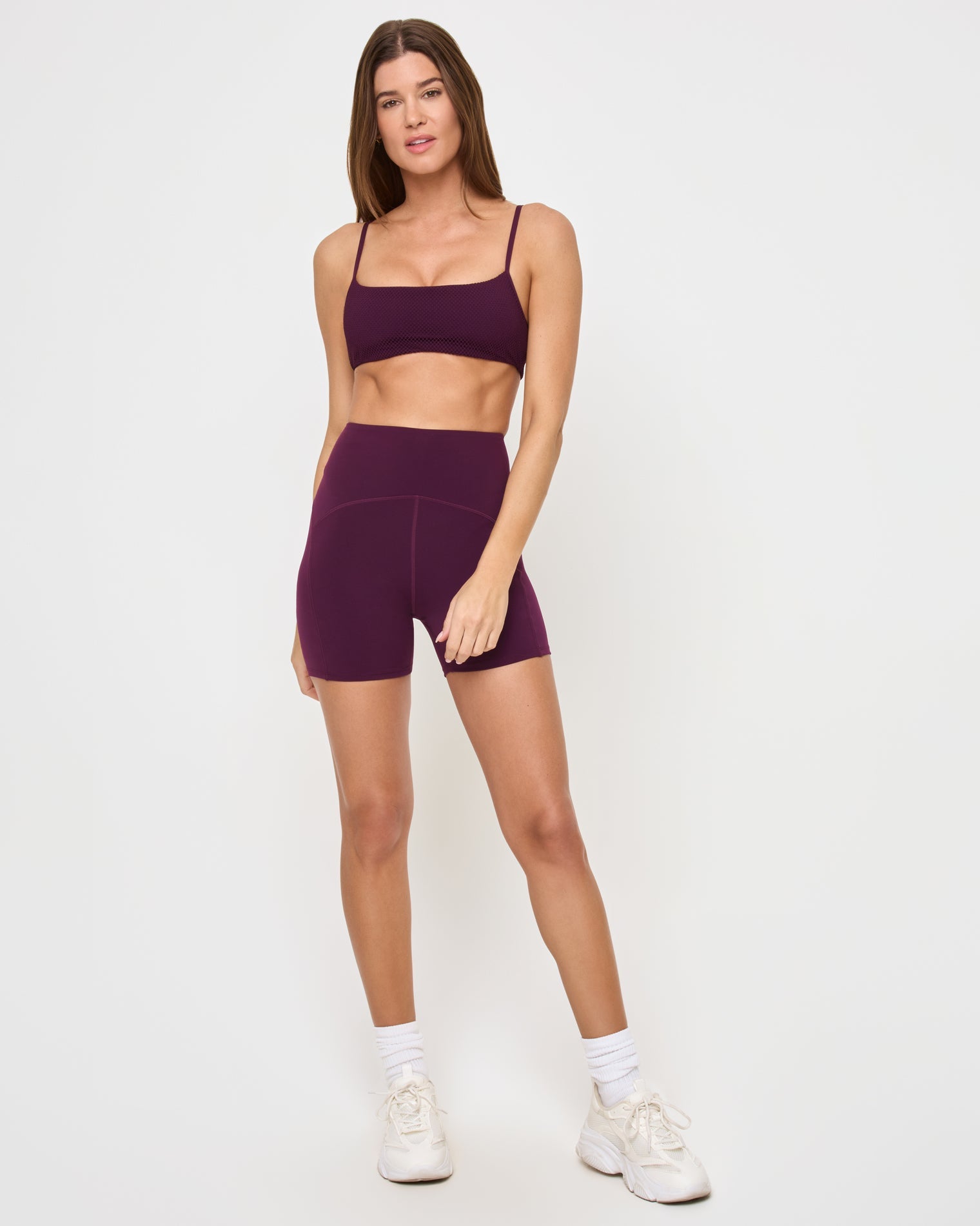 Front view of the LSPACE Stride Bra in plum on a model against a white background, showing the full plum sports bra design, paired with matching plum high-waist active shorts.