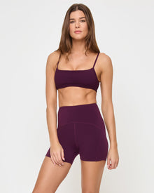 Angled view of the LSPACE En Pointe Short in plum on a model against a white background, showing the profile of the high-waist active shorts, paired with the matching plum Stride Bra.