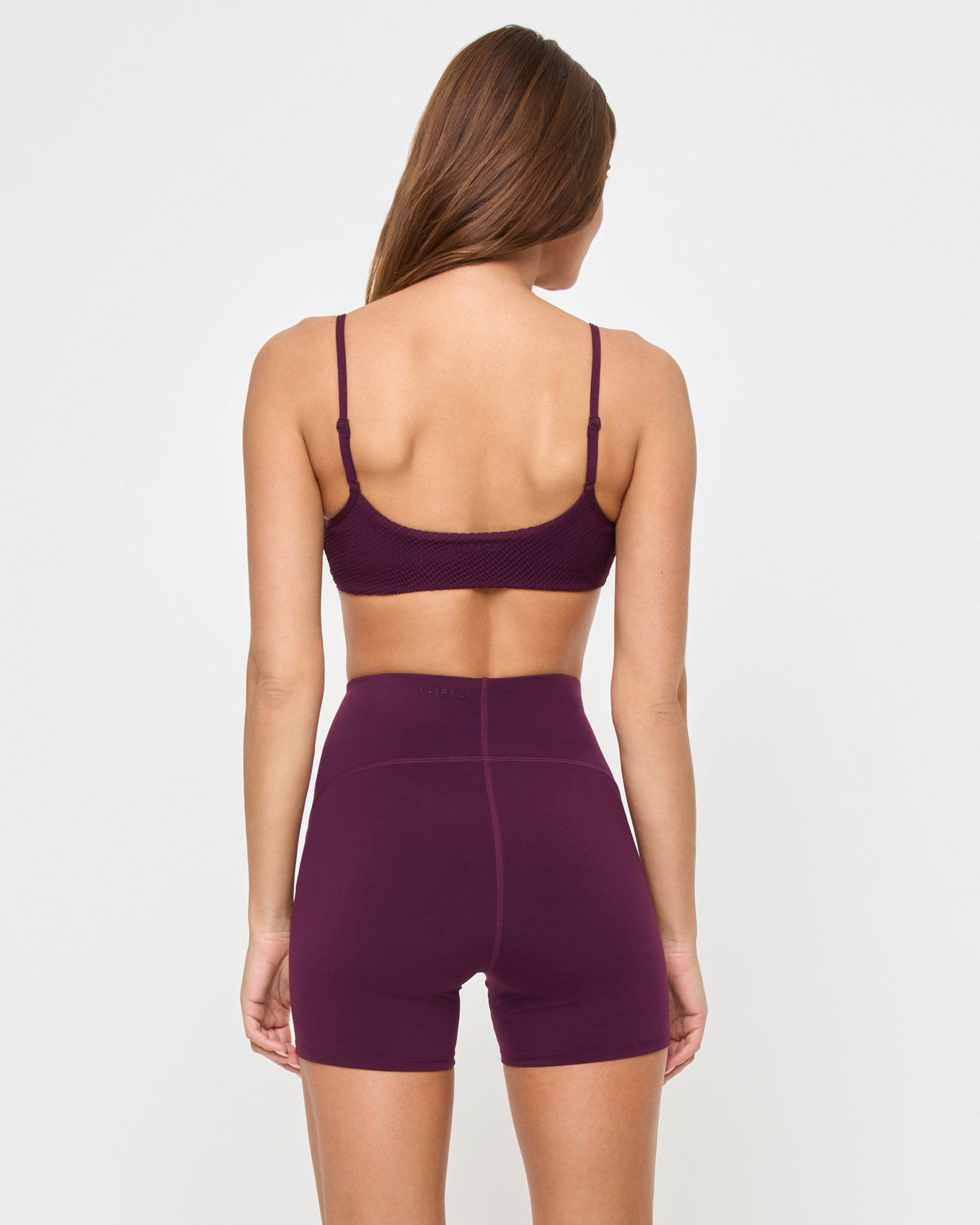 Back view of the LSPACE Stride Bra in plum on a model against a white background, showing the back strap detail of the plum sports bra, paired with matching plum high-waist active shorts.