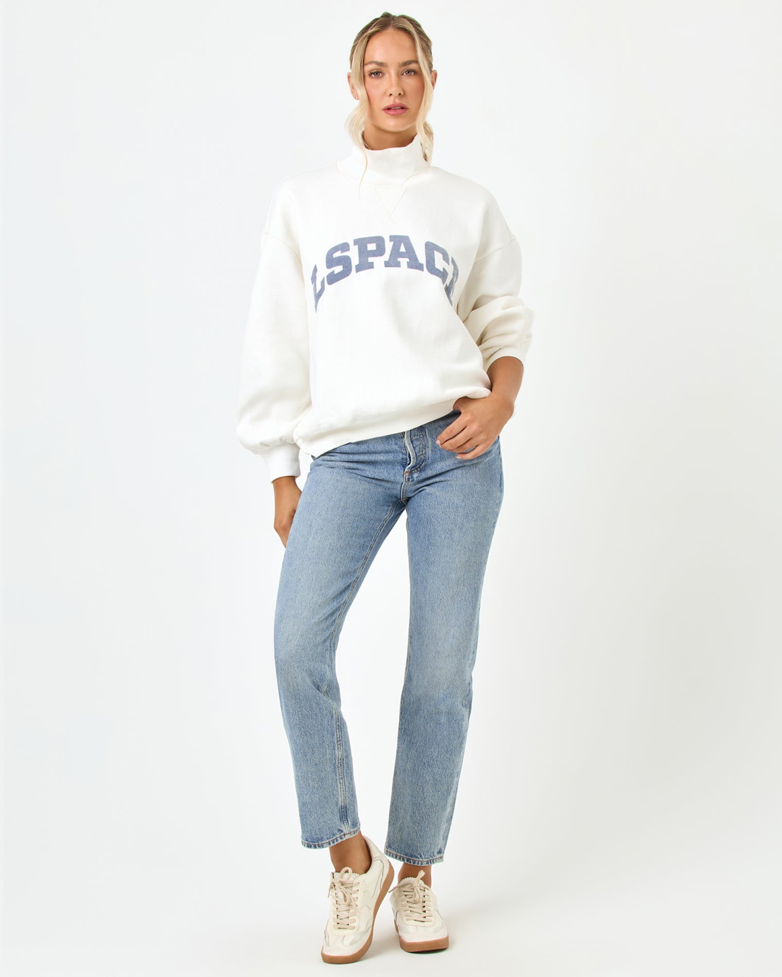 Front full-length studio view of the Crush Sweatshirt in cream, a cream quarter-zip pullover sweatshirt with "LSPACE" collegiate arch lettering in blue and slate, styled with light blue jeans and white sneakers.