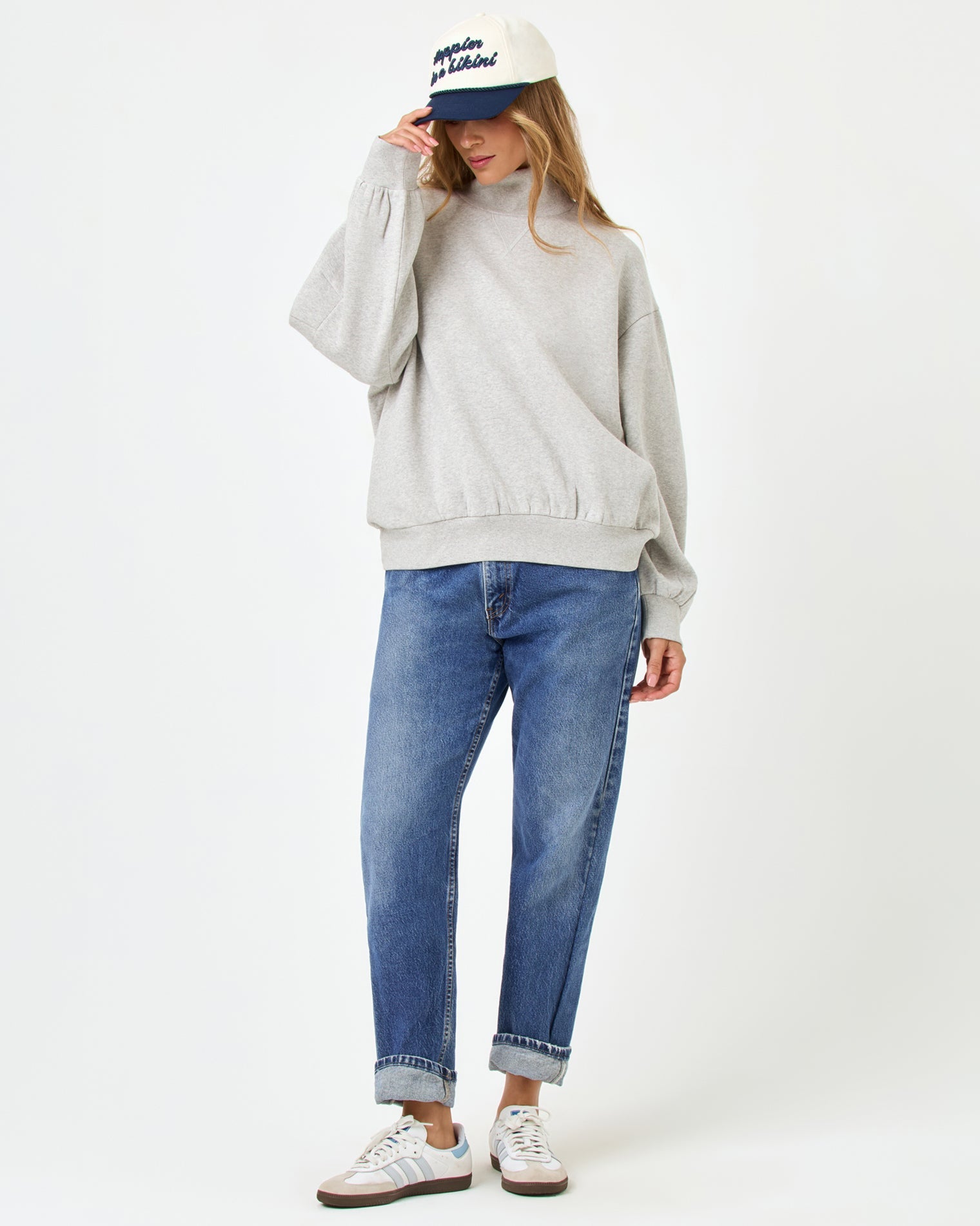 Front full-length studio view of the Crush Sweatshirt in Heather Gray, a heather gray oversized quarter-zip sweatshirt, styled with cuffed blue jeans, Adidas sneakers, and a navy and cream baseball cap.