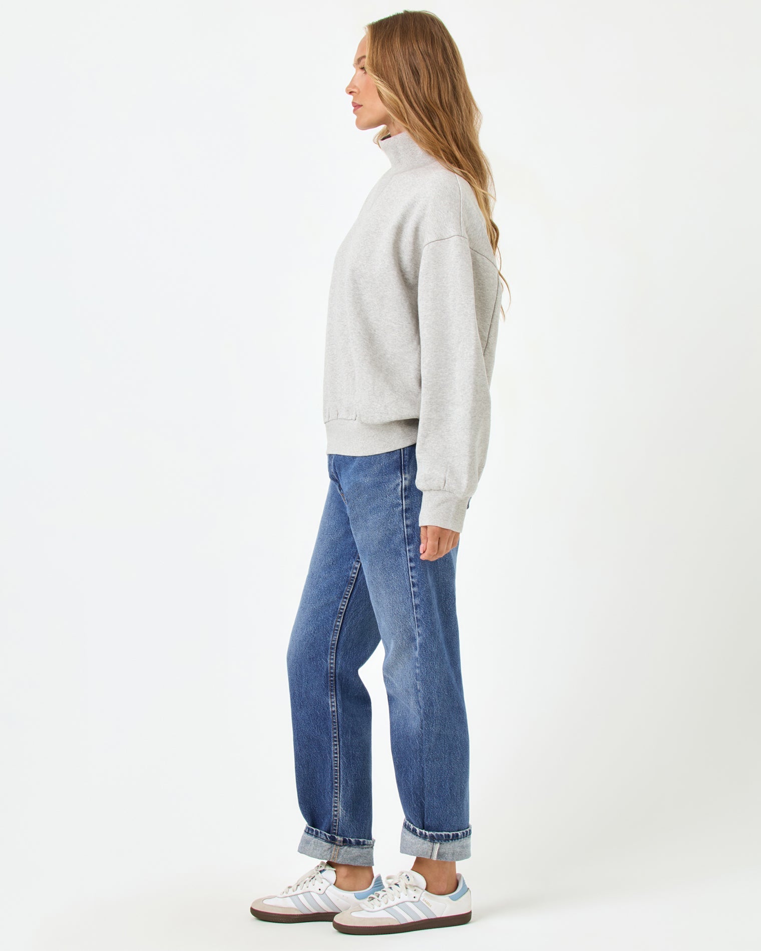 Side profile full-length studio view of the Crush Sweatshirt in Heather Gray, a heather gray oversized quarter-zip sweatshirt, styled with cuffed blue jeans and Adidas sneakers.