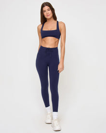 Front view of the LSPACE Uppercut Bra in midnight blue on a model against a white background, showing the full-length outfit with the midnight navy square-neck sports bra and matching high-rise full-length leggings or pants with lace-up waistband, white sneakers and socks.