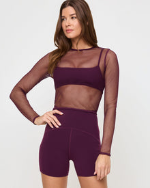 Close-up front view of the LSPACE Stride Bra in plum under a mesh shirt on a model against a white background, paired with plum active shorts, showing an alternate styling option.