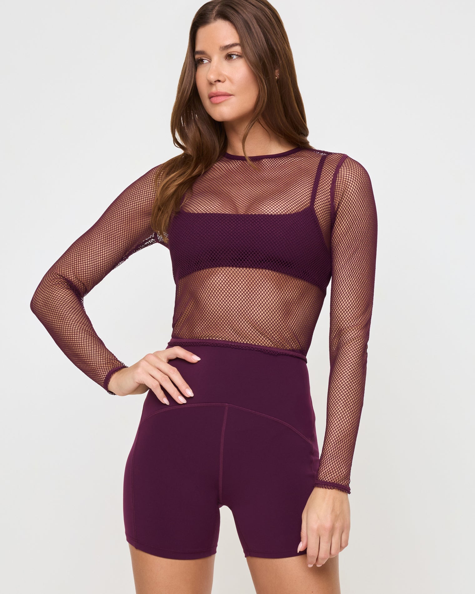 Close-up front view of the LSPACE Stride Bra in plum under a mesh shirt on a model against a white background, paired with plum active shorts, showing an alternate styling option.