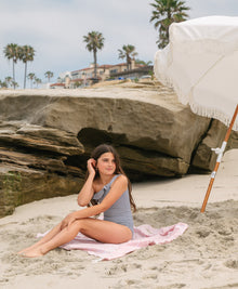 Young girl wearing a striped one piece bikini sitting on a towel on the beach.