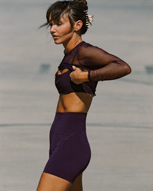 Lifestyle editorial side view of a model wearing the LSPACE En Pointe Short in plum paired with a plum mesh fishnet long-sleeve crop top and biker shorts, in an outdoor sandy setting.