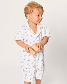 A young child with blonde hair wears the Baby's Twill Summer Romper in Easter Frolic—white with colorful animal prints and yellow trim—standing against a plain light background, holding two wooden blocks and looking to the side.