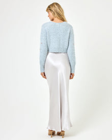 Back view of a model wearing the LSPACE Sweet As Honey Skirt in silver, showing the smooth satin back and A-line silhouette, styled with a pale blue fluffy cardigan and heeled sandals on a white background.