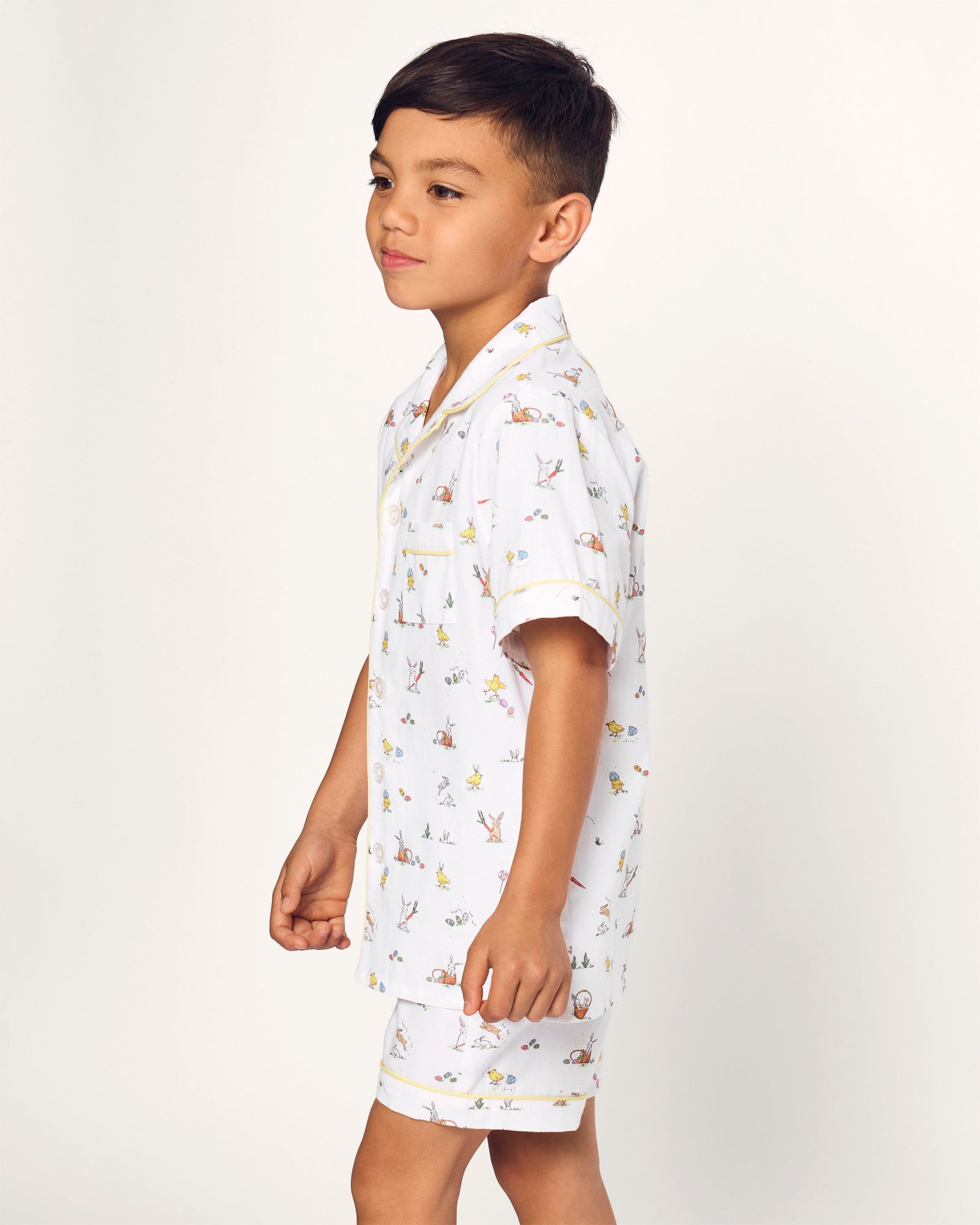 Two smiling children in luxurious Petite Plume sleepwear sit on the floor. They sport matching Kids Twill Short Sets in an Easter Frolic print. One child cuddles a stuffed bunny, embodying comfort and joy against a plain backdrop.