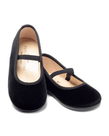 The Kids Delphine Slipper in Black Velvet by Petite Plume features a rounded toe and a thin top strap. One shoe stands upright while the other leans, showing its beige inner lining—ideal for stylish little feet.
