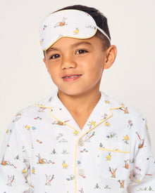 A young boy wears a Kid's Twill Sleep Mask in Easter Frolic, featuring colorful animal and nature prints, along with matching white pajamas. He smiles softly against a plain, light background.