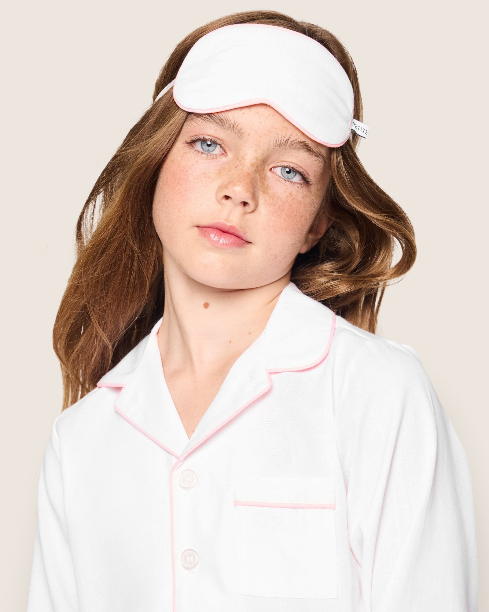 A young girl with long hair and freckles wears a luxurious white pajama top with pink trim and a matching monogrammed Kids Twill Sleep Mask in White with Pink Piping by Petite Plume. She gazes forward against a plain light beige background.