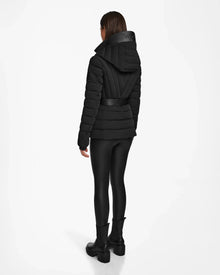 A person stands facing away, wearing the Rudsak ALISA - L124506 Black womens quilted down puffer jacket with a high collar, black leggings, and black boots against a plain white background.