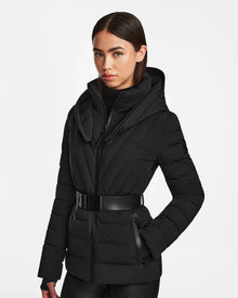 A woman with straight dark hair wears the Rudsak ALISA - L124506 Black, a womens quilted down puffer with vegan leather belt, high collar, and hood. She stands against a white background with hands in pockets and a neutral expression.