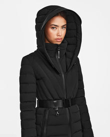 A woman in the Rudsak ALISA - L124506 Black quilted down puffer coat with a vegan leather belt stands against a plain white background, looking directly at the camera with a neutral expression.