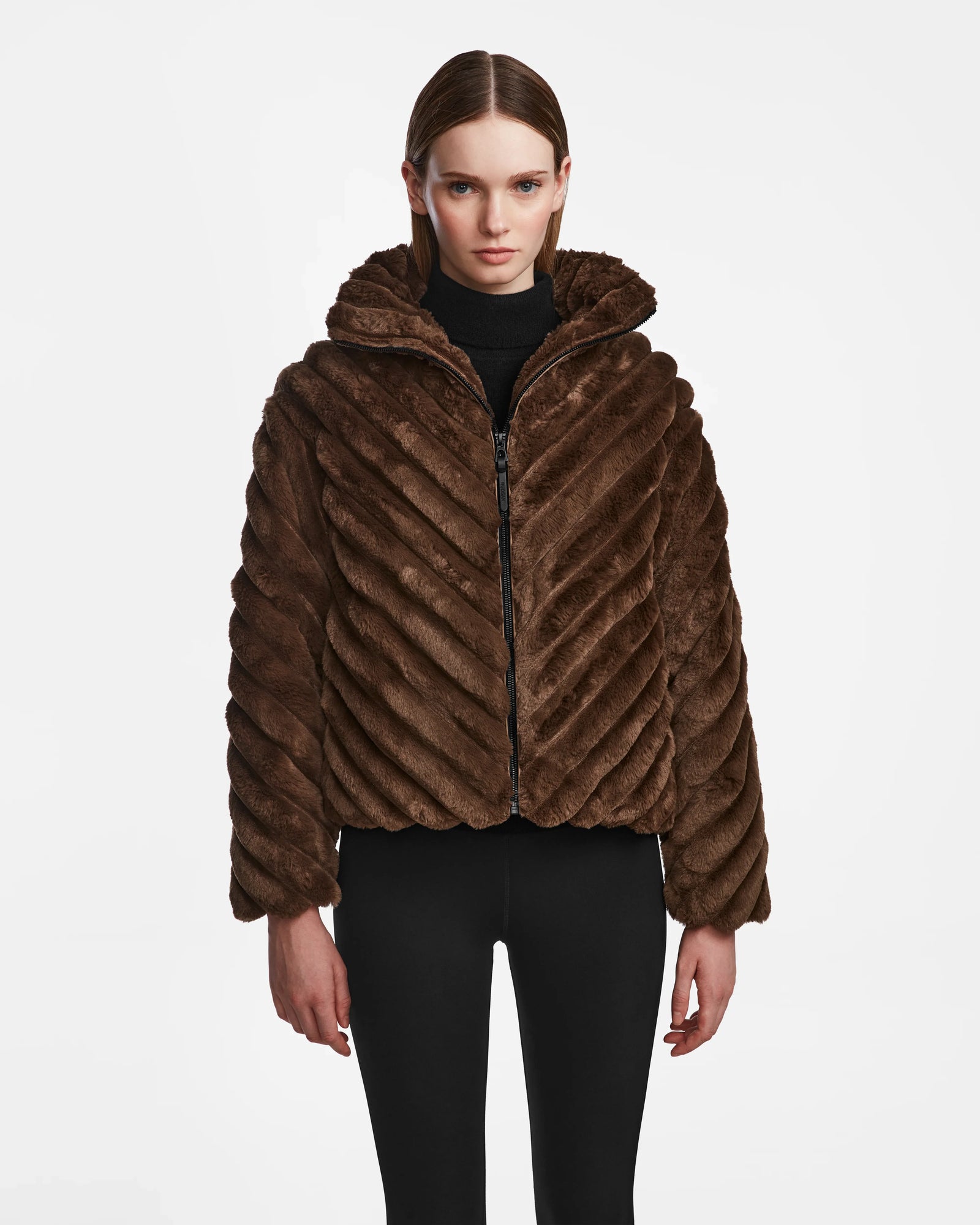 A woman faces forward wearing the Rudsak ANASTASIA - 8124804 Espresso, a textured brown faux fur zip-up jacket with a hood and black fitted pants, styled against a plain white background.