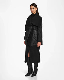 A woman stands against a white background wearing the Rudsak ANNE MARIE - 8124713 Black quilted down wool jacket with large lapels, paired with a black turtleneck, black front-slit midi skirt, and black knee-high boots.