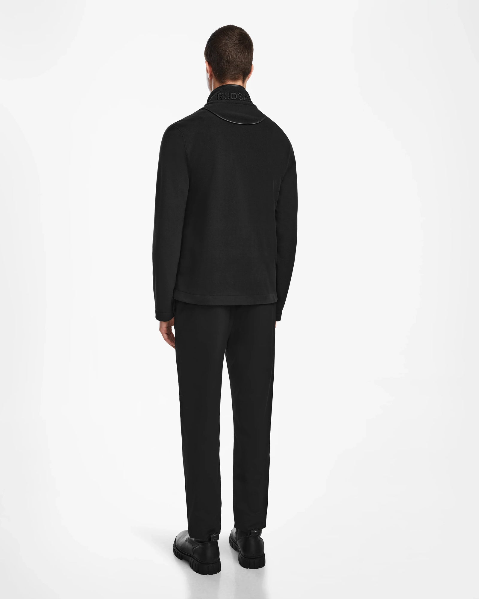 A man faces away from the camera, wearing the Rudsak ARAGON - 6524539 Black mens fleece jacket with vegan leather trims, paired with black pants and shoes against a plain white background.