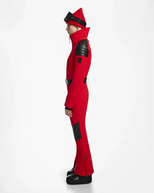 A person in profile wears the Rudsak ARLEY - 8124717 Flame Red fitted one-piece ski suit with black padded details, a matching red beanie, black ski goggles on the forehead, and black boots against a plain white background.