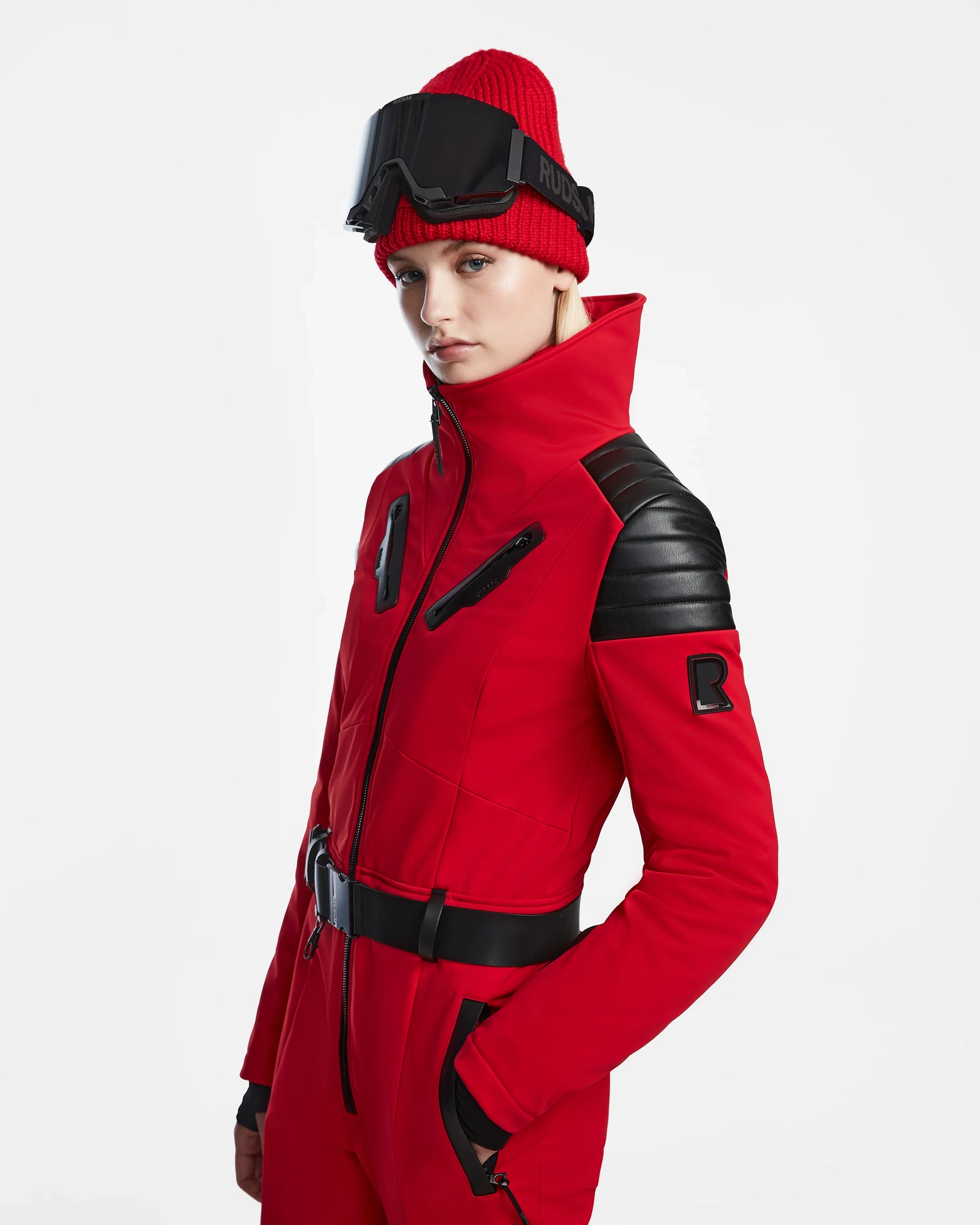 A person wears the Rudsak ARLEY - 8124717 Flame Red fitted one-piece ski suit with black accents, a red knit beanie, and black ski goggles on their forehead, standing against a plain white background.