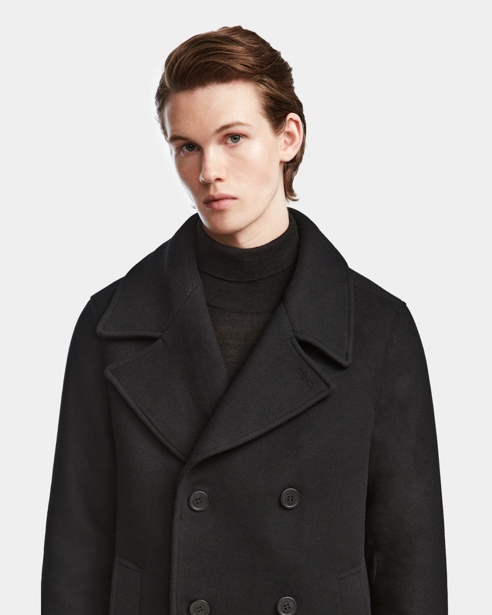 A young person with fair skin and light brown hair, wearing the Rudsak ARTHUR - 6124521 Black premium wool blend double-breasted peacoat over a black turtleneck, stands against a plain white background, facing slightly right.