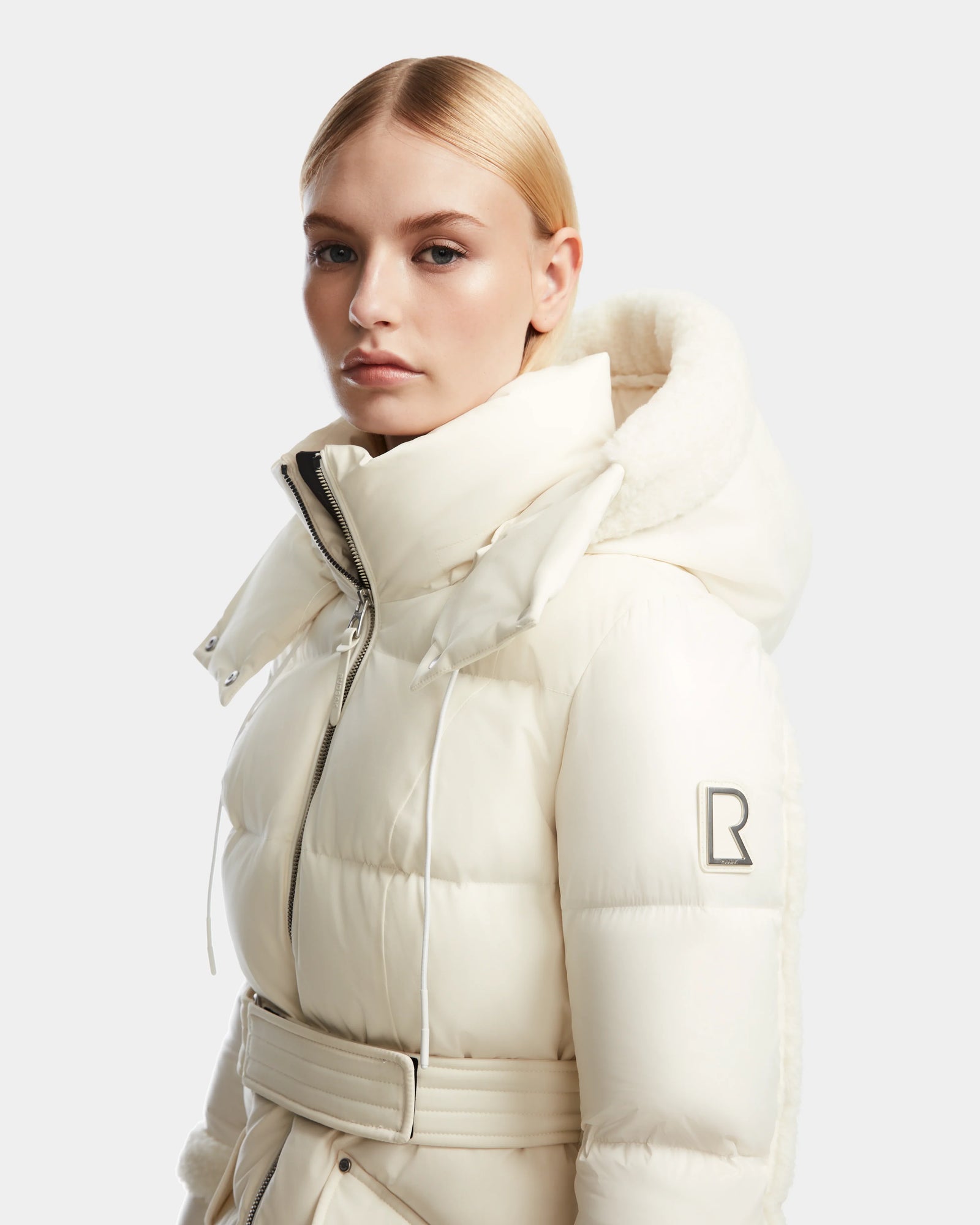 A blonde woman in a sleek low part wears the off-white Rudsak AUDREY L - 8124585 Iceland knee-length down puffer with high collar, hood, RDS certified down, and B logo patch on the sleeve, posing against a plain background.