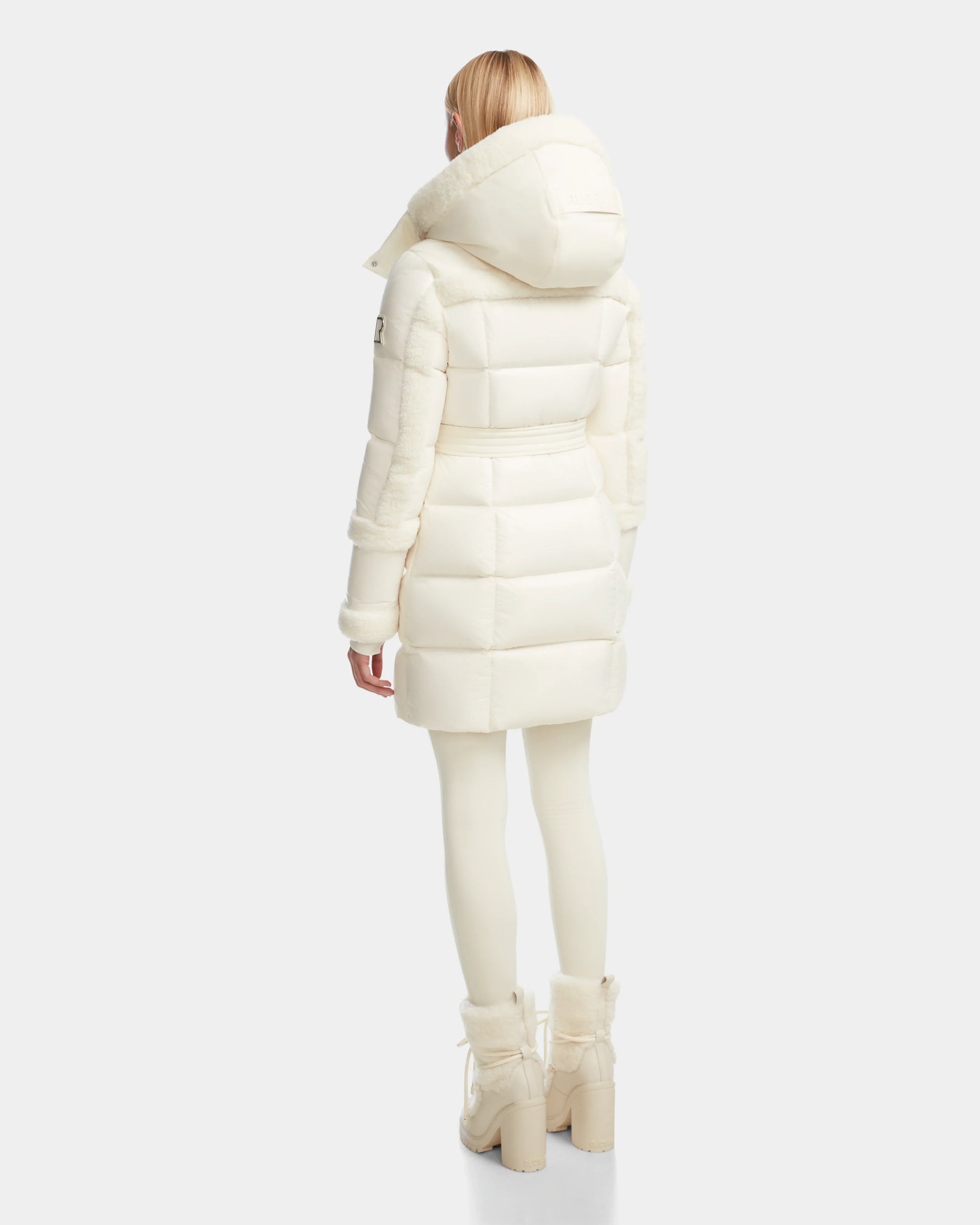 A person with blonde hair is wearing the Rudsak AUDREY L - 8124585 Iceland knee-length down puffer jacket, matching leggings, and heeled boots, standing and facing away on a light gray background.