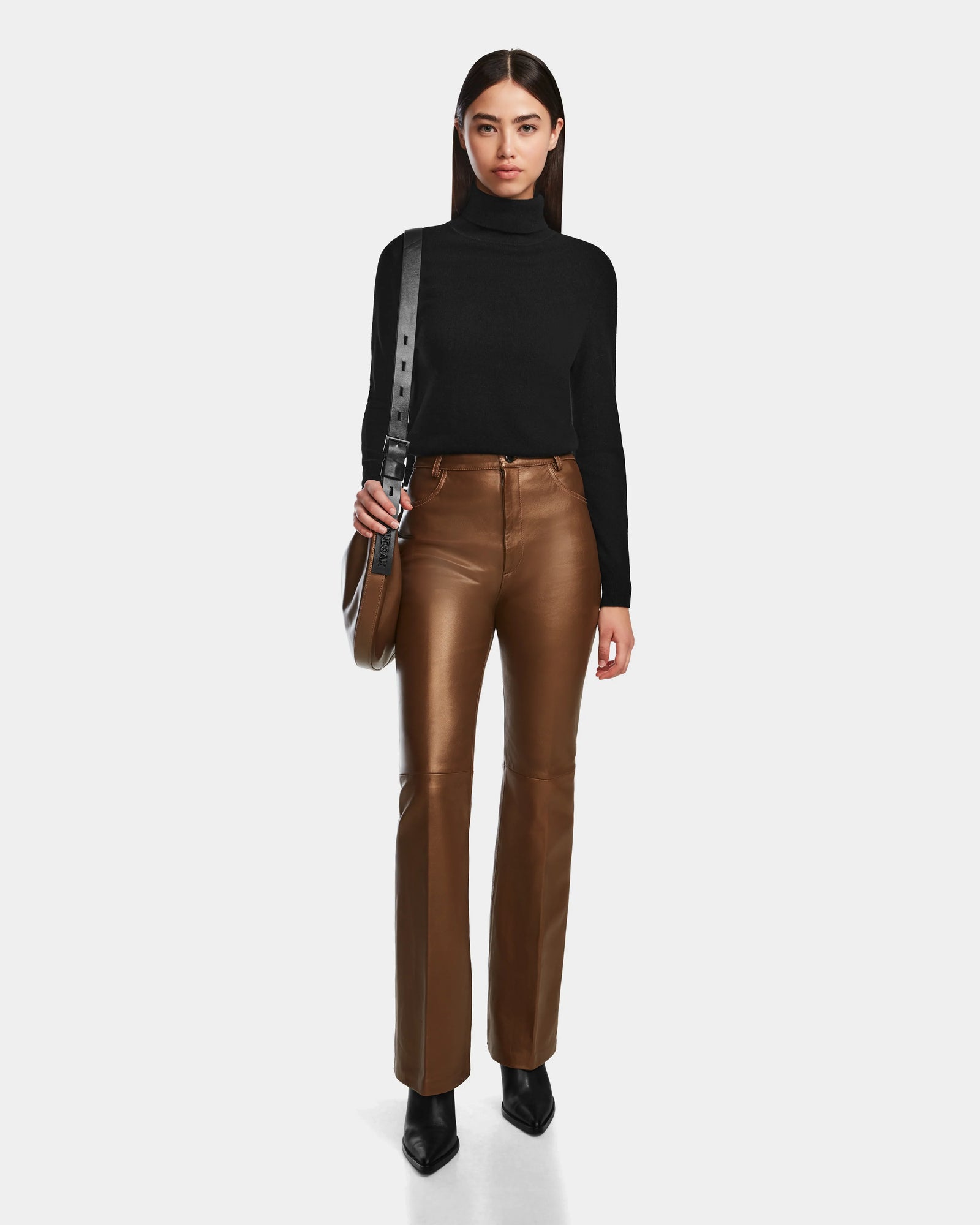 A woman stands facing forward in the Rudsak CARISSA - 8524545 Antique Bronze leather pants, black turtleneck, pointed black shoes, and a black shoulder bag against a plain white background.