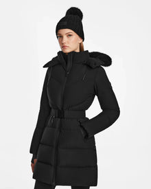 A woman wears the Rudsak CHRISTEL M-M - L124505 Black quilted winter coat with a belted waist, paired with a black knit pom-pom beanie and black pants, standing against a plain white background.