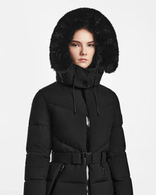 A woman wearing the Rudsak CHRISTELLE M - L123525 Black sustainable winter coat with a fitted silhouette and large fur-lined hood stands against a white background, looking straight ahead with a neutral expression.