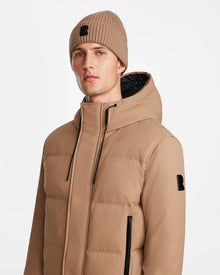 A man wears the Rudsak DALLAS - M122501 Sand weather-resistant bomber jacket with hood and black zippers, paired with a matching tan beanie, looking to the side against a plain white background.