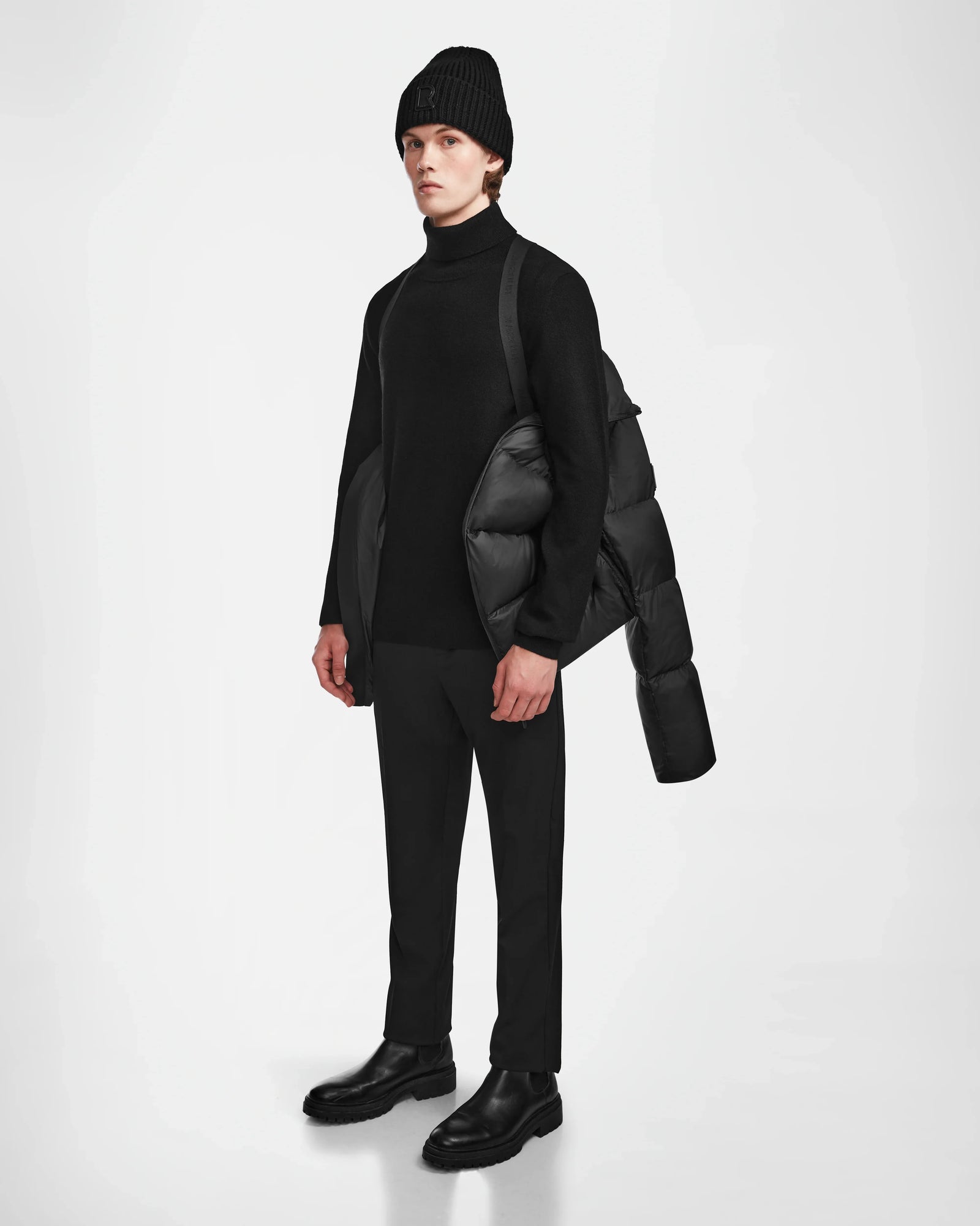 A person stands against a white background in an all-black outfit with the Rudsak DANIEL M - M124510 Black weather-resistant jacket draped over one shoulder.